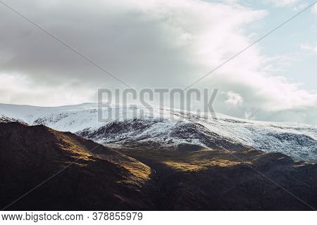 Atmospheric View To Snowy Mountains In Sunlight Under Cloudy Sky. Low Clouds Among Big Rocks With Sn
