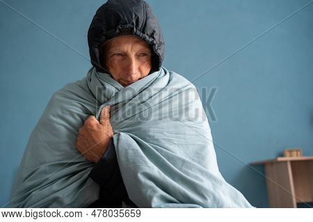 Senior Man Freezing At Home In Winter, No Heating. Elderly Person Wearing Hat And Wrapped In Blanket