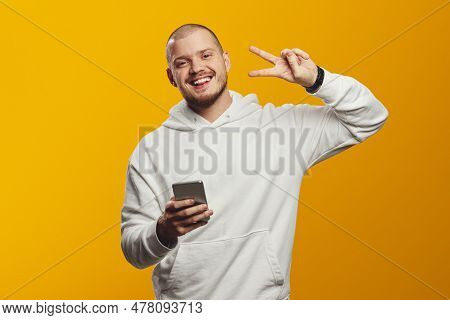 Excited Man Wearing White Hoodie, Smiling And Showing V Sign While Using Cellphone Against Yellow Ba