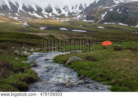 Orange Tent Near Clear Water Stream With View To Mountain Range Among Low Rainy Clouds. Dramatic Alp