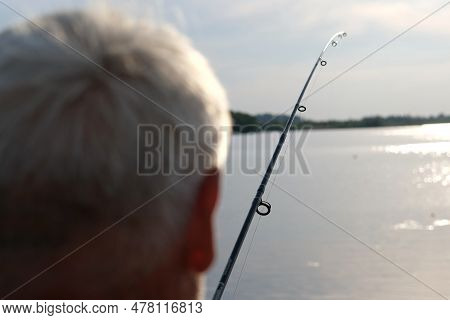 Old Man Fishing. Senior Gray Haired Fisherman Throws A Spinning From Shoreside At Sunset, Twisting A
