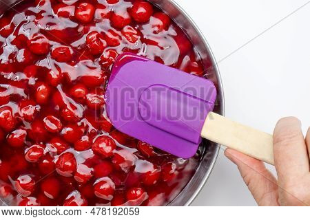 A Hand Holding A Purple Rubber Spatula Spreading Cherry Pie Filling In A Silver Pan. Clean White Tab