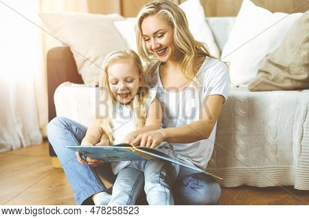 Happy Family. Blonde Young Mother Reading A Book To Her Cute Daughter While Sitting At Wooden Floor 