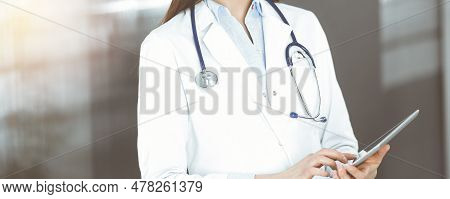 Unknown Woman Doctor Iusing Tablet Computer In Her Hands, While Standing In Sunny Clinic. Medicine C