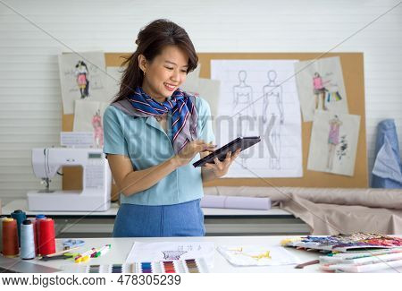 Young Asian Dressmaker In Striped Scarf Checking Email Order From Tablet Computer. Pattern For Cloth