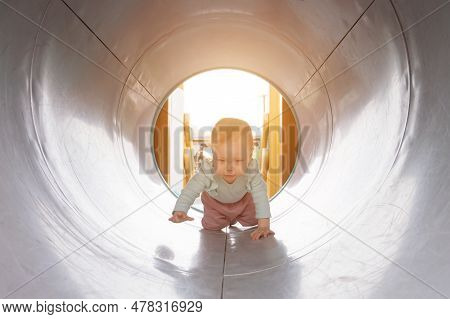 Toddler Girl Likes To Crawl Along Gray Pipe On Kids Playground. Daughter With Excited And Amused Exp