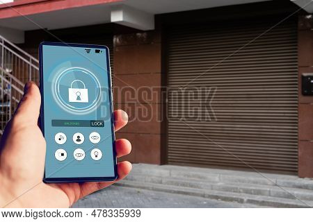 Female Hand Holding Black Mobile Smart Phone With Smart Home Application On The Screen. Blurred Hous