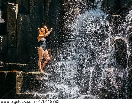 Caucasian Woman In A Black Swimsuit Refreshing Under Falling Water Streams Flowing On Black Volcanic