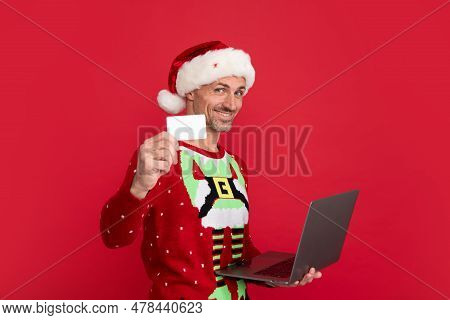 Santa With Credit Card And Laptop. Portrait Of Middle Aged Man In Sweater Isolated Over Red Backgrou