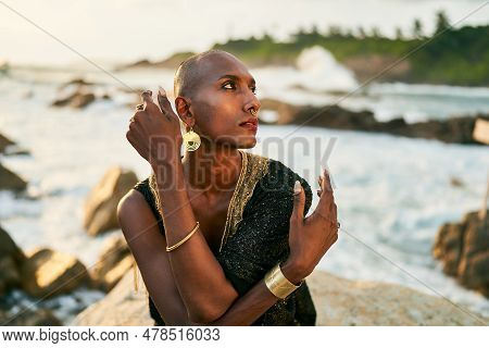 Non-binary Black Person In Luxury Dress Sits On Rocks In Ocean. Trans Ethnic Fashion Model Wearing J