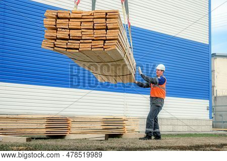 A Slinger Unloads Wooden Planks Into A Pile. A Worker In A Helmet And A High-visibility Vest Unloads