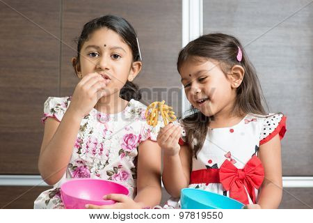 Two cute Indian girls eating food. Asian sibling or children enjoying traditional snack murukku, living lifestyle at home.