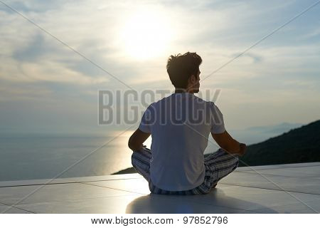 handsome young man practicing yoga on in modern home terace with ocean and sunset in background
