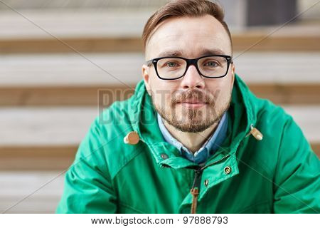 people, style, leisure and lifestyle - happy young hipster man in eyeglasses and jacket sitting on stairs in city