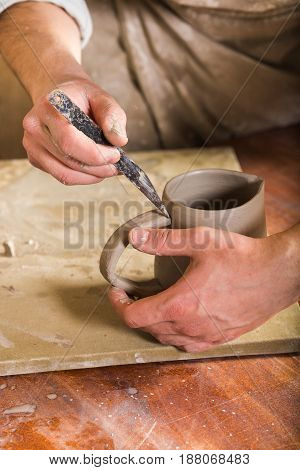pottery, stoneware, ceramics art concept - closeup of craftsman hands connect handle to cup with tool, master fingers work with pieces of fireclay, male stand at workshop behind table, vertical