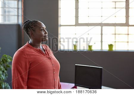Focused young African businesswoman looking deep in thought while standing alone by windows in a large modern office