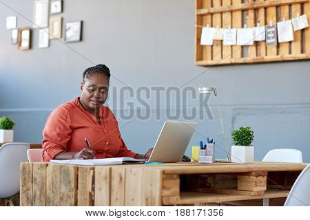 Young African businesswoman working on a laptop and writing down notes in a book while working at her desk in a large modern office
