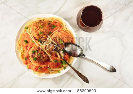 Overhead photo of plate of meatballs with pasta, tomato sauce, fresh parsley, shot from above on white marble texture with place for text, with fork and spoon plunged into dish, with glass of red wine