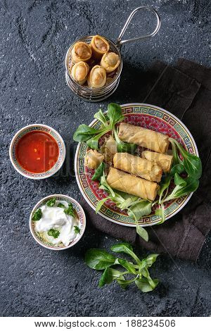 Fried spring rolls with red and white sauces, served in traditional china plate and fry basket with fresh green salad over black texture background. Flat lay, space. Asian food