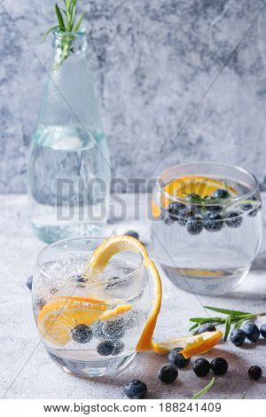 Tonic water cocktail with rosemary, blueberries and orange. Two cold glasses and bottle with zest and bubbles over gray blue texture background. Refreshing beverage alco non alcohol