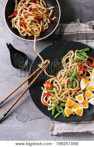 Cooking stir fry udon noodles, green beans, sliced paprika, boiled eggs, soy sauce with sesame seeds in black plate with wood chopsticks over gray texture background. Flat lay. Asian style dinner