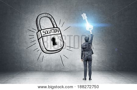 Rear view of a businessman in a dark suit holding a giant key standing near a black wall with a large padlock with a success word on it. Toned image