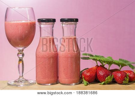 A glass and two bottles with strawberry smoothie on a pink background