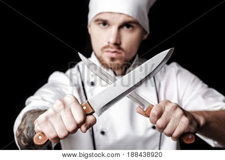 Young bearded man chef In white uniform holds Two knives on a black background. Focus on knives