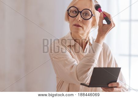 Do make up. Elegant elderly woman keeping brush in right hand and wrinkling forehead while looking straight at camera