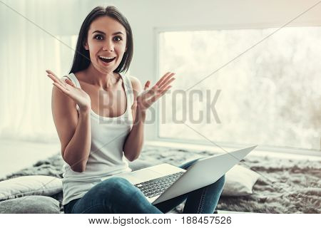 Happy young woman is working with a laptop at home looking at camera and showing surprise
