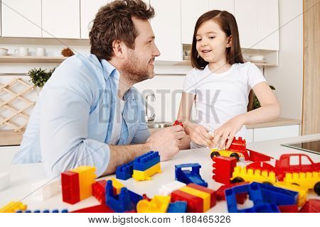 Lovely day together. Young bristled man smiling at his daughter and holding a red construction set part while she trying to assemble a car model staying near the surface full of toy pieces