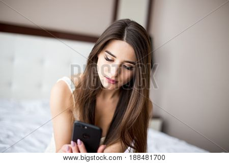 A Woman Using Her Smartphone As She Sitting Onthe Bed And Smiling.