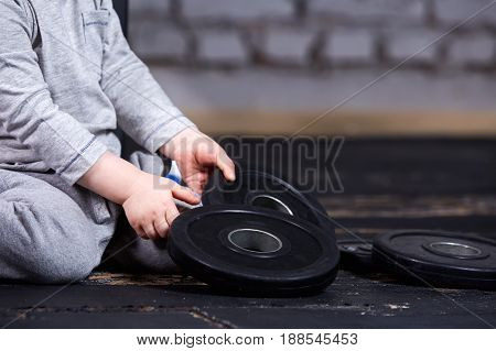 Close-up view photo of little sporty child with barbbell against brick wall at the cross fit gym. Little boy in the grey sportwear. Rearing children in the healthy lifestyle.