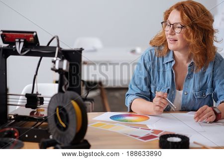 At work. Nice smart hard working woman sitting at the table and looking at the 3D printer while doing some drawings