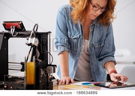 Designer studio. Intelligent female 3d designer standing near the table and leaning at it while working on a tablet