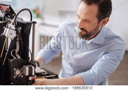 New design. Handsome professional male designer looking at the 3d printer and creating a new design while working with 3d technology
