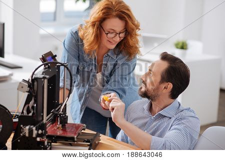 Let me look. Happy pleasant nice woman leaning over the table and taking a small 3d printed object while wanting to look at it