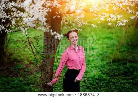 Young Beautiful Girl In A Pink Shirt Standing Under Blossoming Apple Tree And Enjoying A Sunny Day.