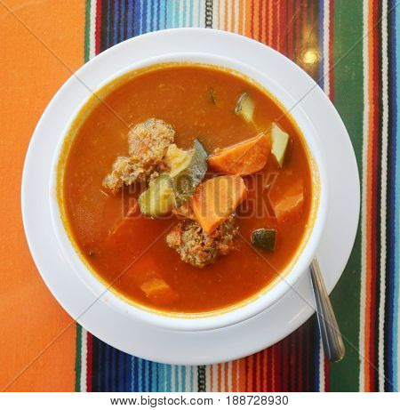 Traditional Mexican food mexican soup with meat balls and vegetables. Mexican  albondigas soup in a white ceramic bowl and plate on a traditional Mexican serape blanked table cloth and a spoon. 