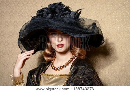 Portrait of a gorgeous young woman in elegant broad-brimmed hat and luxurious dress over golden background. Vintage style. Beauty, fashion.