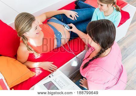 Youg girl watching midwife attaching CTG to pregnant belly of mother-to-be
