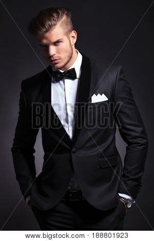 elegant young fashion man in tuxedo with half shaded face and hands in  pockets is looking at the camera.on black background