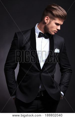 elegant young fashion man in tuxedo with his hands in his pockets looking away from the camera, down to his side.on black background