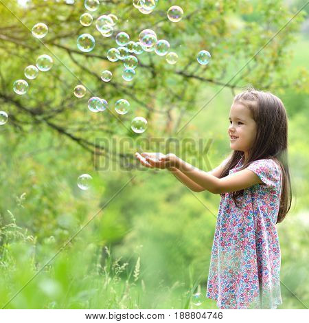 Cute little girl on a forest glade among lush vegetation and around fly soap bubbles. Kid with soap bubbles over nature outdoor.