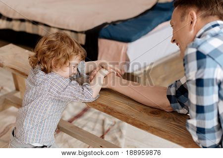 Be winner. Attractive kid with curly hair putting both hands on the palm of his father while sitting in semi position near bench and looking downwards