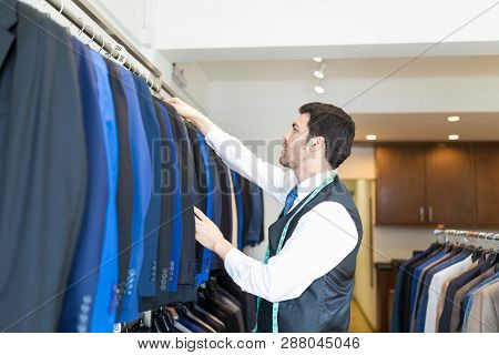Side View Of Good Looking Male Seamster Arranging Suits In Rack At Clothing Showroom