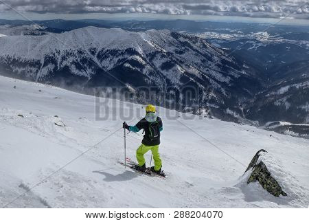 A Man Skier Finding The Best Track For Freeride. A Skier Looking Down To The Valley. A Yellow Helmet