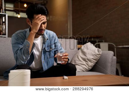 Illness And Unhealthy Condition Concept. Asian Man Sitting On Sofa Having Headache Holding Glass Of 