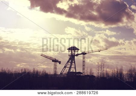 Silhouettes Of Old Coal Mine Tower And Two Cranes Against Dramatic Skyscape In Genk, Belgium. Indust