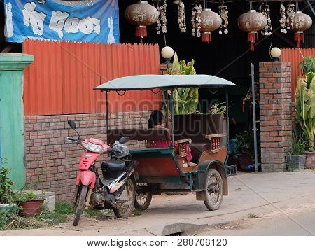 Cambodia, Siem Reap 12/08/2018 A Little Asian Girl Sits In A Moto Rickshaw Near A House With Red Lan